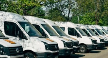 A large fleet of white vans parked in neat rows, stretching across a spacious lot. The vans are uniform in color and size, with clean exteriors reflecting the bright sunlight. Some have company logos, while others remain plain, all ready for deliveries or transport.